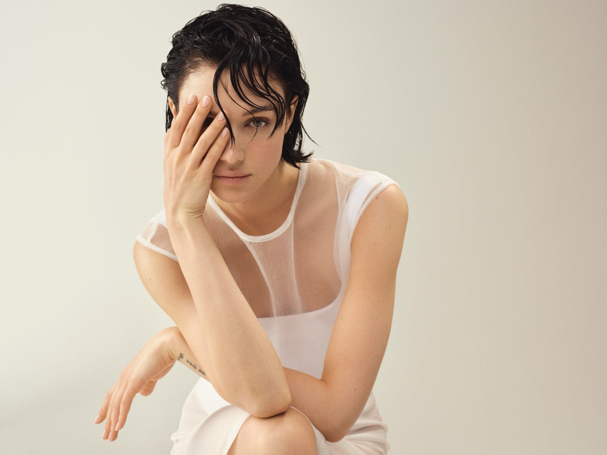 Modelo con cabello corto oscuro y efecto húmedo cubriéndose un ojo con la mano, fondo crema