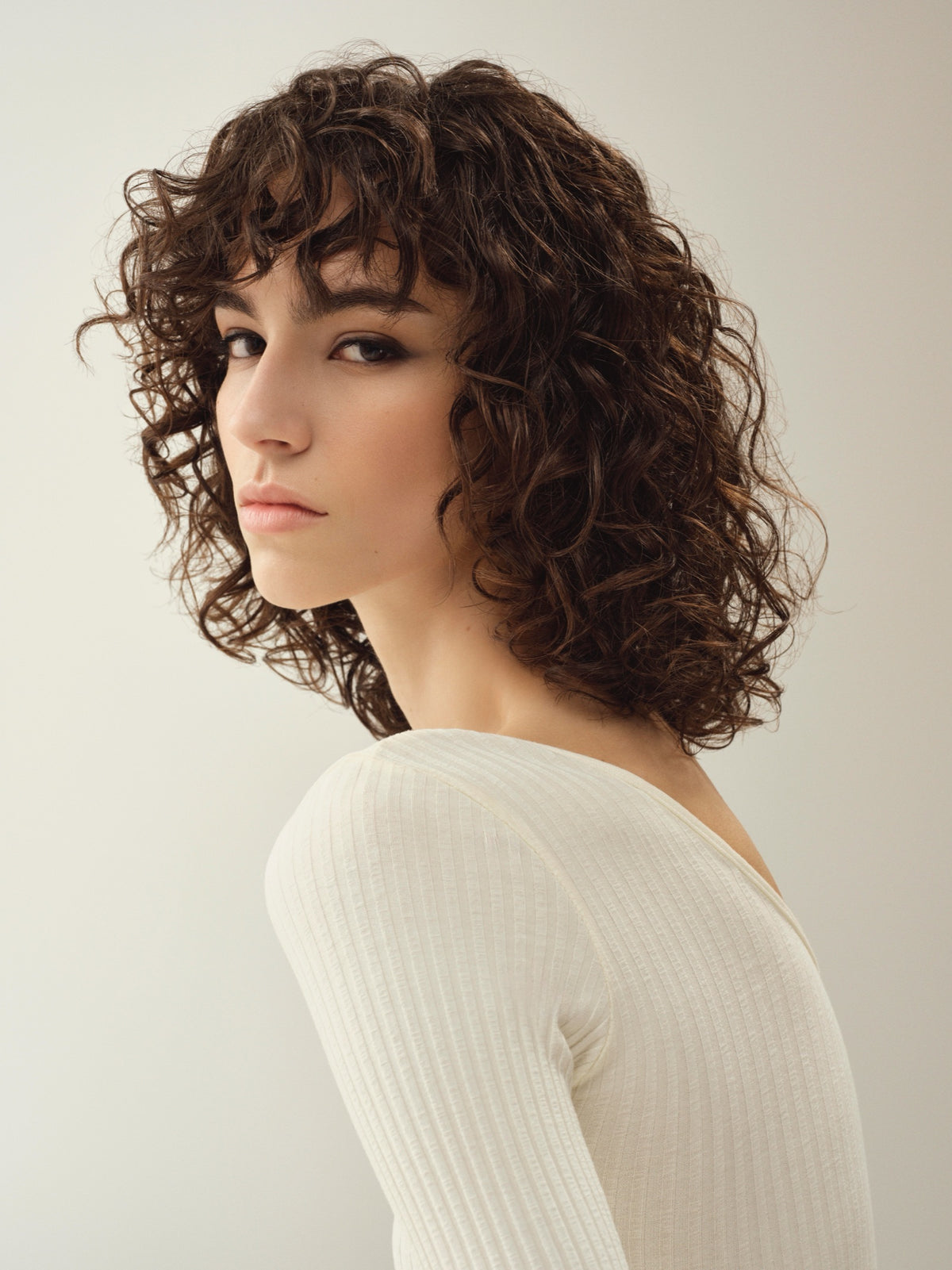 Modelo de cabello rizado castaño oscuro con suéter color marfil sobre fondo crema